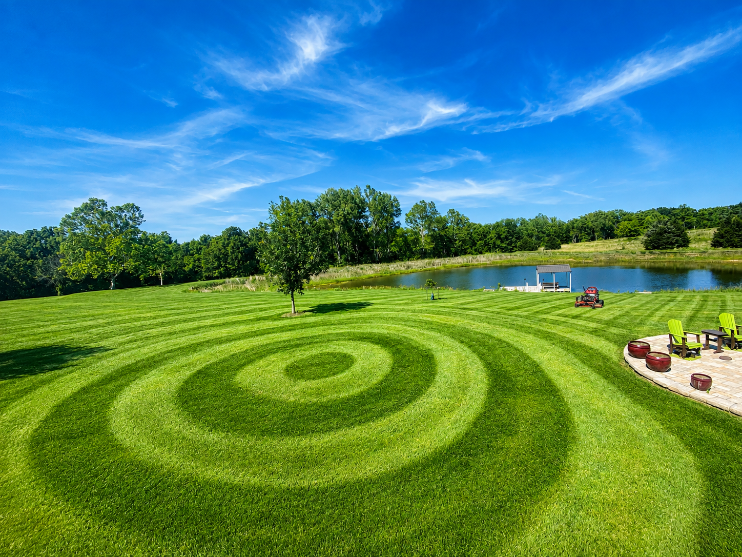 A large lawn mowed in concentric circular stripes beside a pond, with a riding mower parked near the patio.